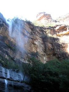 _spray from a waterfall, red layered rocks above in the sunlight and green bushy trees in the shadows below_