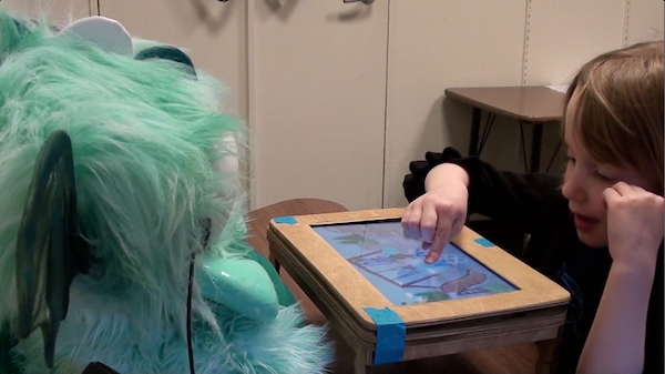 Girl moving a picture on a tablet screen, with the tablet inset in a mini table that is between her and a dragon robot